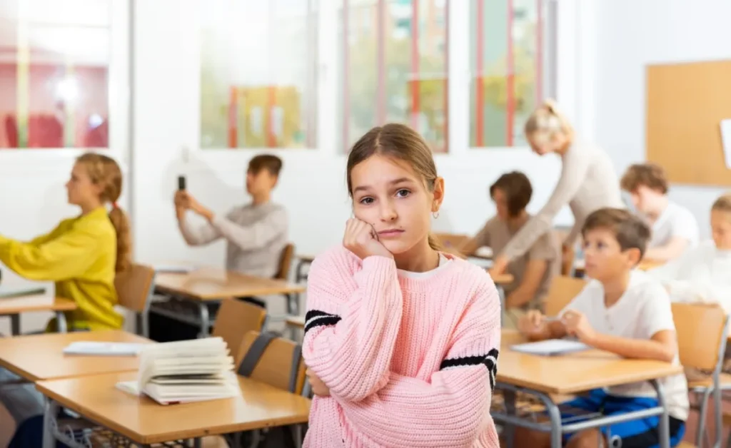 A young girl in a pink sweater sits in a classroom, resting her face on her hand with a bored or disinterested expression, while other students and a teacher engage in various activities in the background.